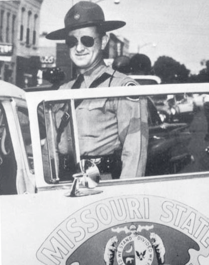 Black-and-white photo of Tpr. Allman in uniform and sunglasses standing beside a Missouri State Highway Patrol vehicle with visible door emblem.