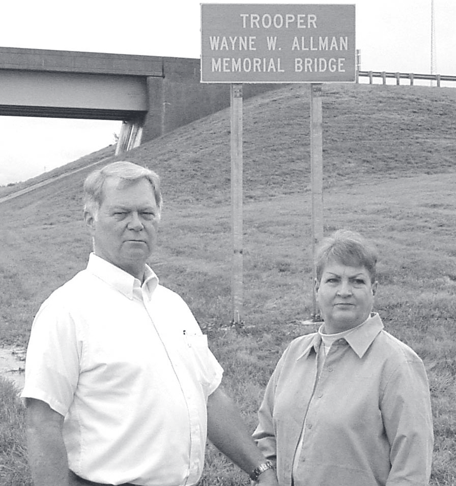 A man and woman stand in front of a highway sign reading 'Trooper Wayne W. Allman Memorial Bridge' with an overpass in the background.
