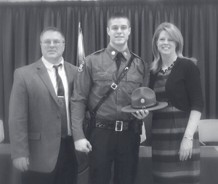 Tpr. Bava in Patrol uniform holds his campaign hat while standing between a man and woman in front of flags.