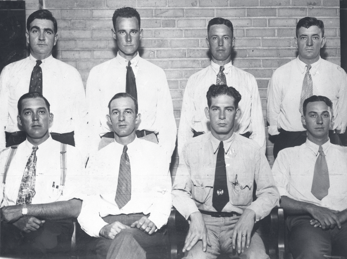 Vintage black-and-white group portrait of eight young men in white shirts and ties, arranged in two rows against a brick wall.