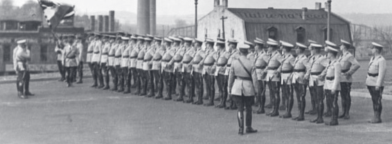 Vintage black-and-white photograph of uniformed officers standing at attention in formation on pavement, with industrial buildings in the background.