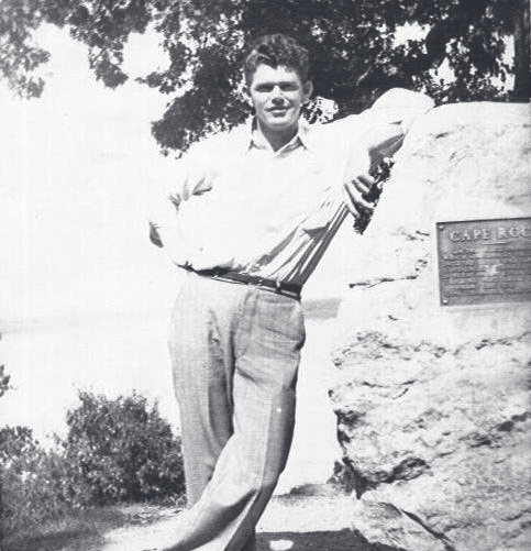 Tpr. Corbin stands with arms crossed beside a large rock bearing a plaque, with water and trees in the background.