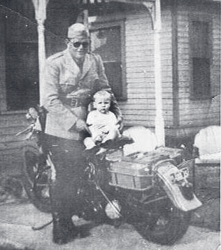Tpr. Corbin in uniform sits on a Patrol motorcycle with a small child in his lap outside a house.