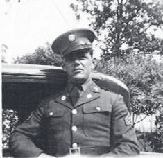 Tpr. Corbin in uniform and service cap stands in front of a dark-colored vehicle with trees behind him.