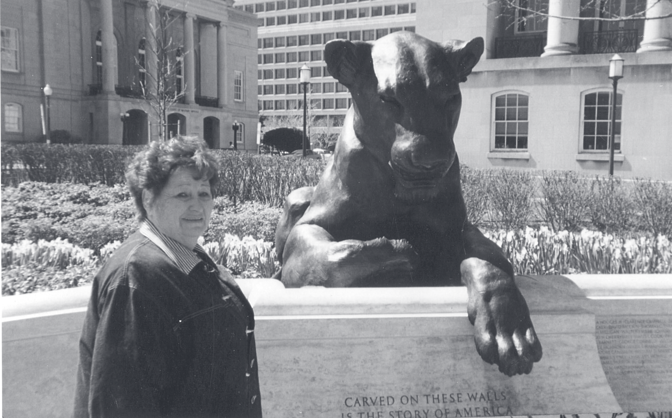 A woman stands beside a bronze lion sculpture mounted on a stone base inscribed 'Carved on these walls is the story of America.'