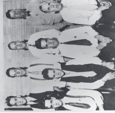 Vintage black-and-white group portrait of eight men arranged in two rows, wearing suits and ties.