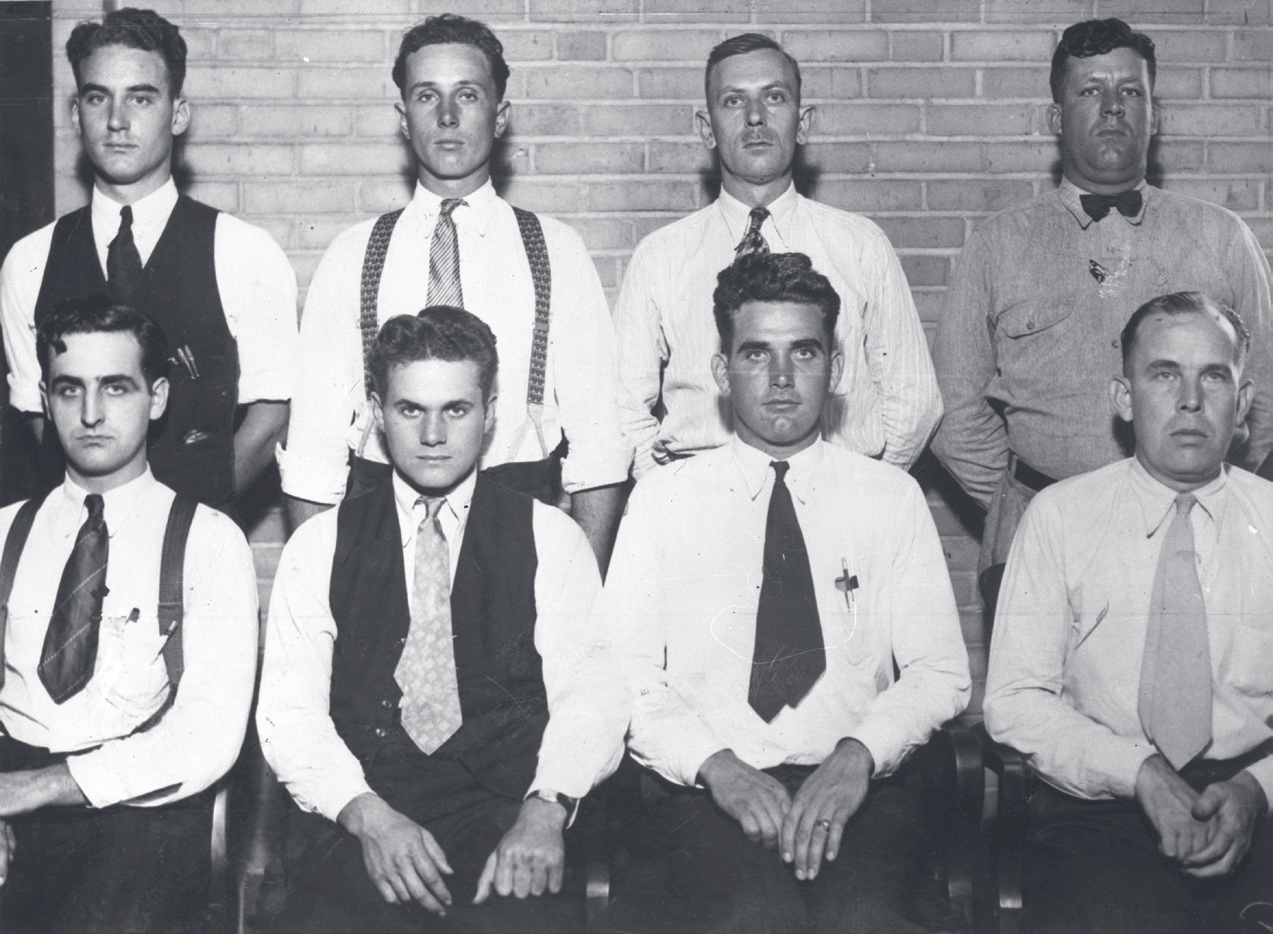 Vintage black-and-white group portrait of eight men in shirts, ties, and vests posed in two rows against a brick wall.