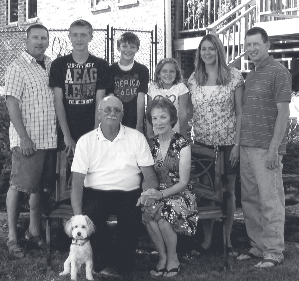 Family members pose together outdoors in front of a brick home with a small white dog seated in front.