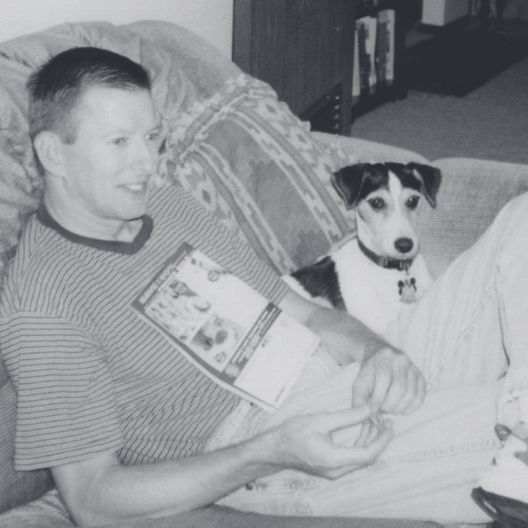 Cpl. Engelhard sits on a couch holding an open magazine next to a small white and brown terrier.