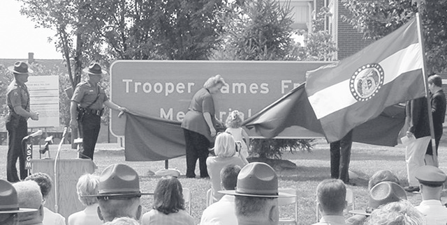 Officers in campaign hats stand at attention beside a draped memorial sign reading 'Trooper James' during an outdoor dedication ceremony.