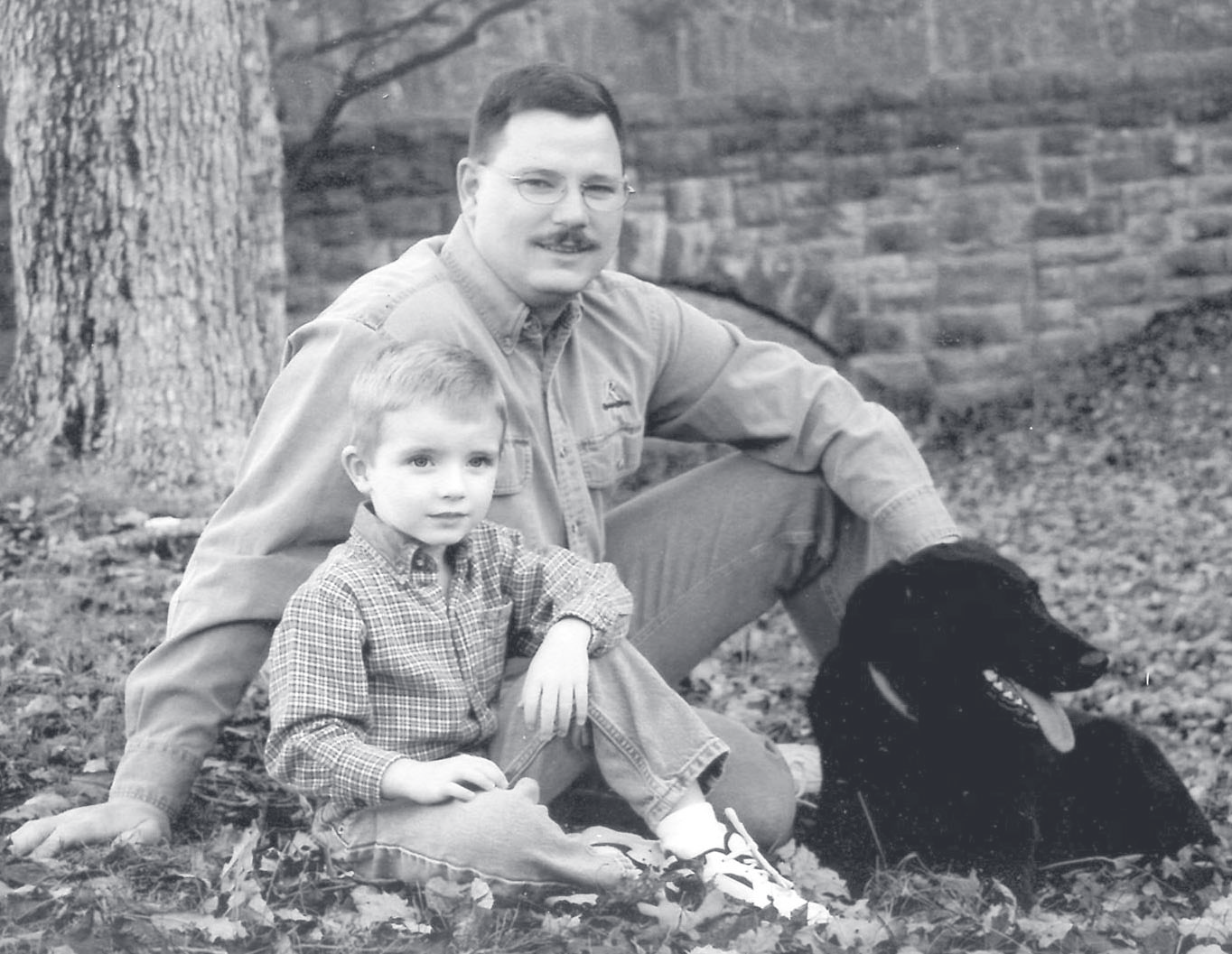 Sgt. Graham sits outdoors among fallen leaves with a young child and a black dog beside a stone wall.