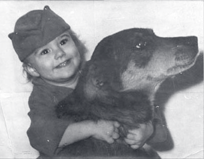 Vintage black-and-white photo of a smiling young child in a soft cap hugging a large dog.