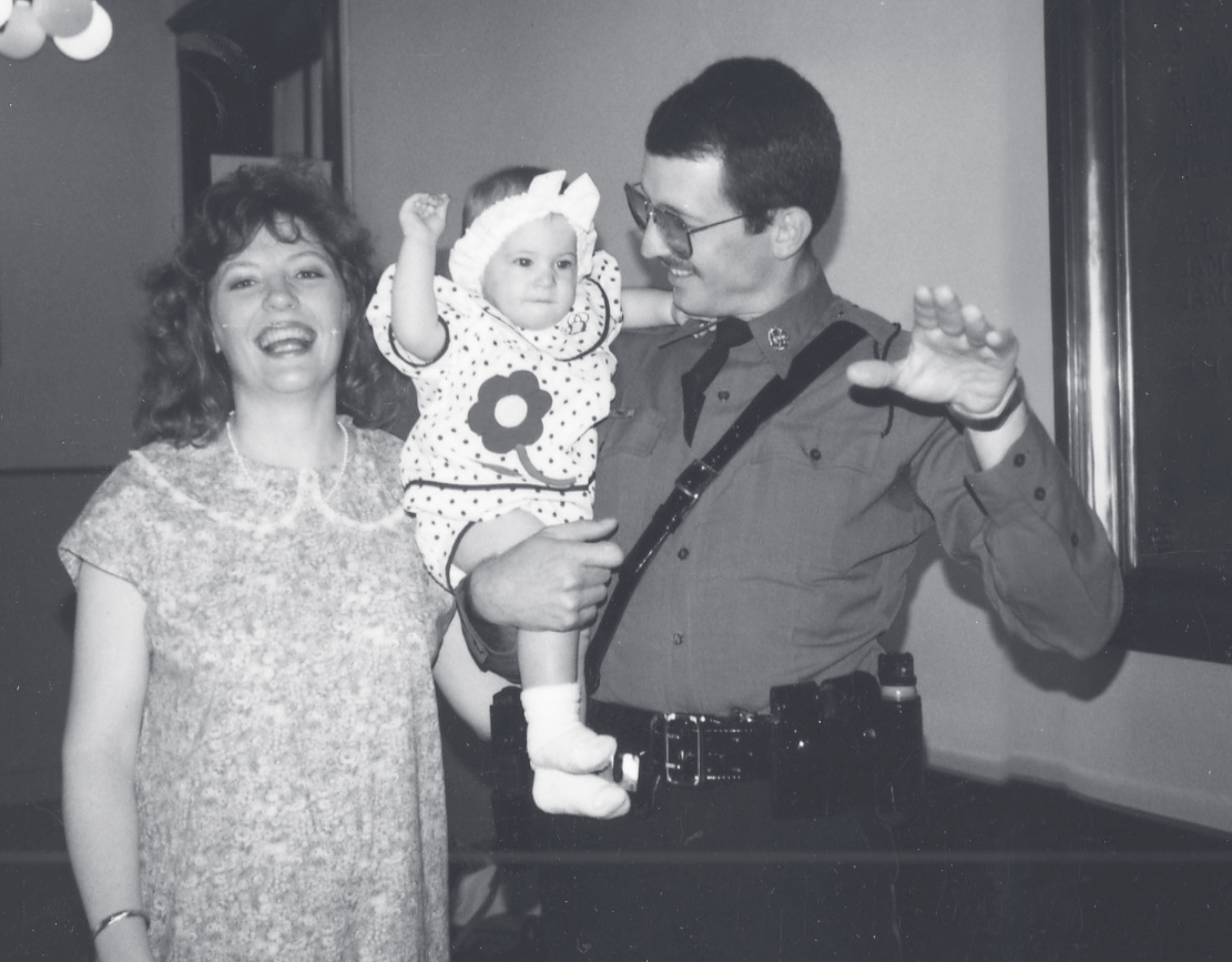 Sgt. Guilliams in uniform holds a smiling toddler beside a laughing woman indoors.