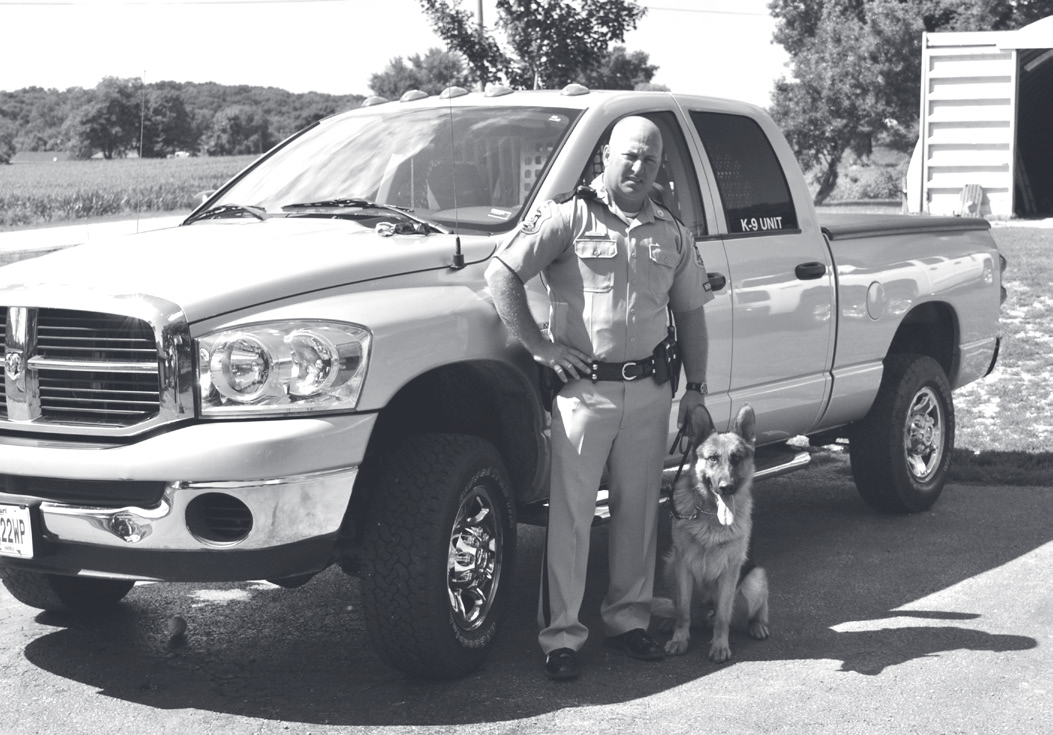 Tpr. Guthrie stands in uniform beside a pickup truck with his German Shepherd K-9 partner sitting at his feet.