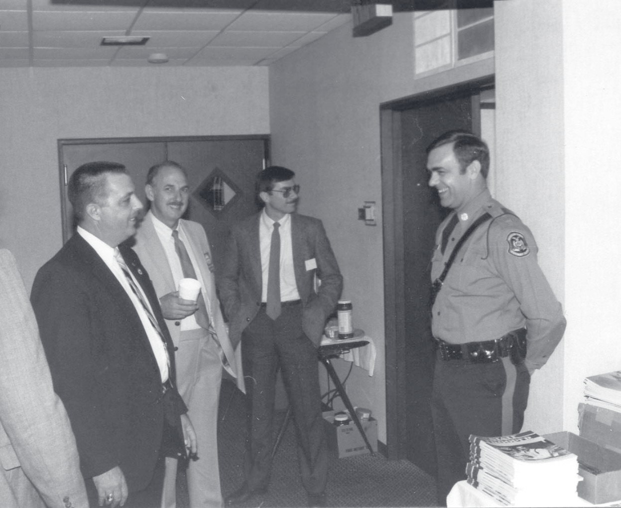 Tpr. Harper in uniform stands in a doorway speaking with three men in business attire holding coffee cups.