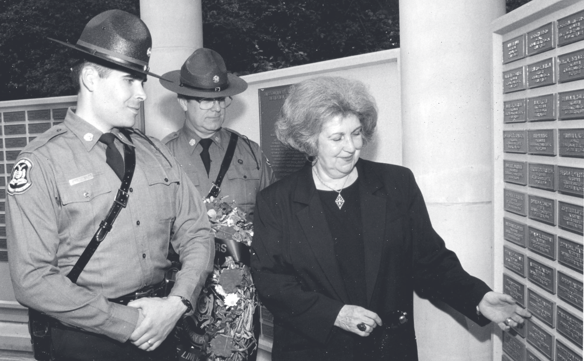 Tpr. Harper and another trooper holding flowers stand with a woman touching an engraved memorial plaque on a columned wall.