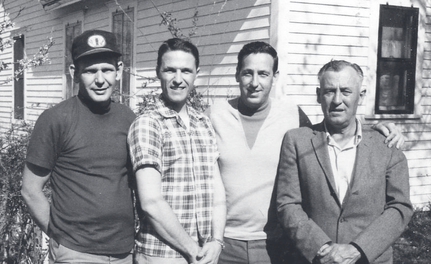 Four men stand in a row outside a white house on a sunny day.