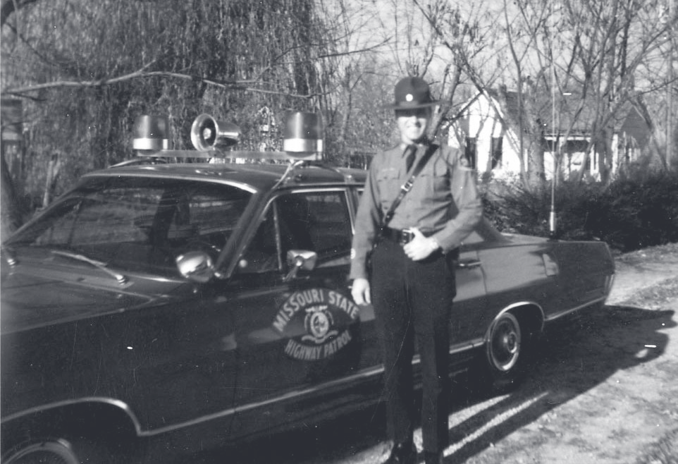 Tpr. Jenkins stands in uniform beside a Missouri State Highway Patrol cruiser in a residential driveway.