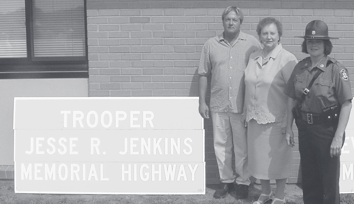 Two family members stand beside a uniformed officer next to a Trooper Jesse R. Jenkins Memorial Highway sign.