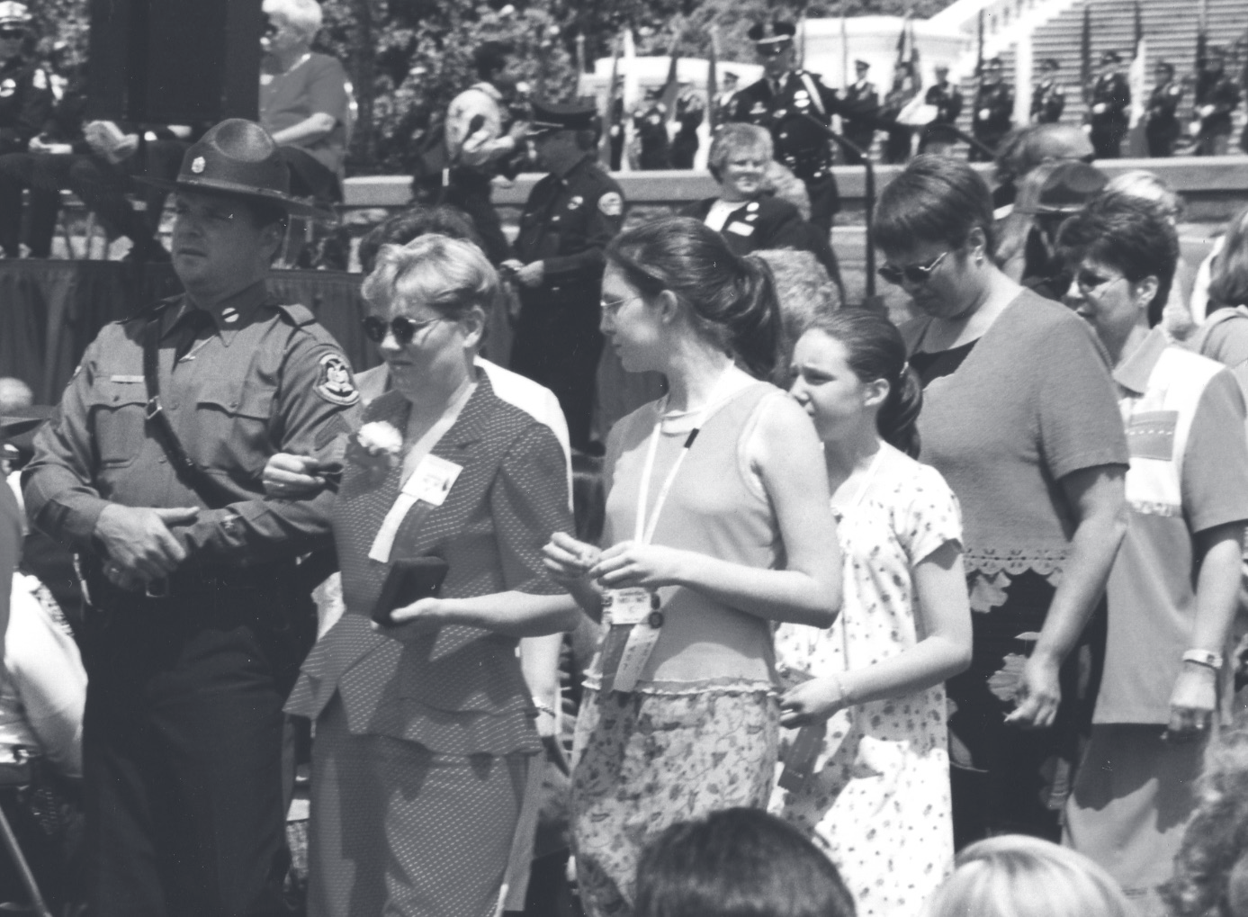 Family members walk alongside a uniformed trooper at an outdoor memorial ceremony with officers in the background.