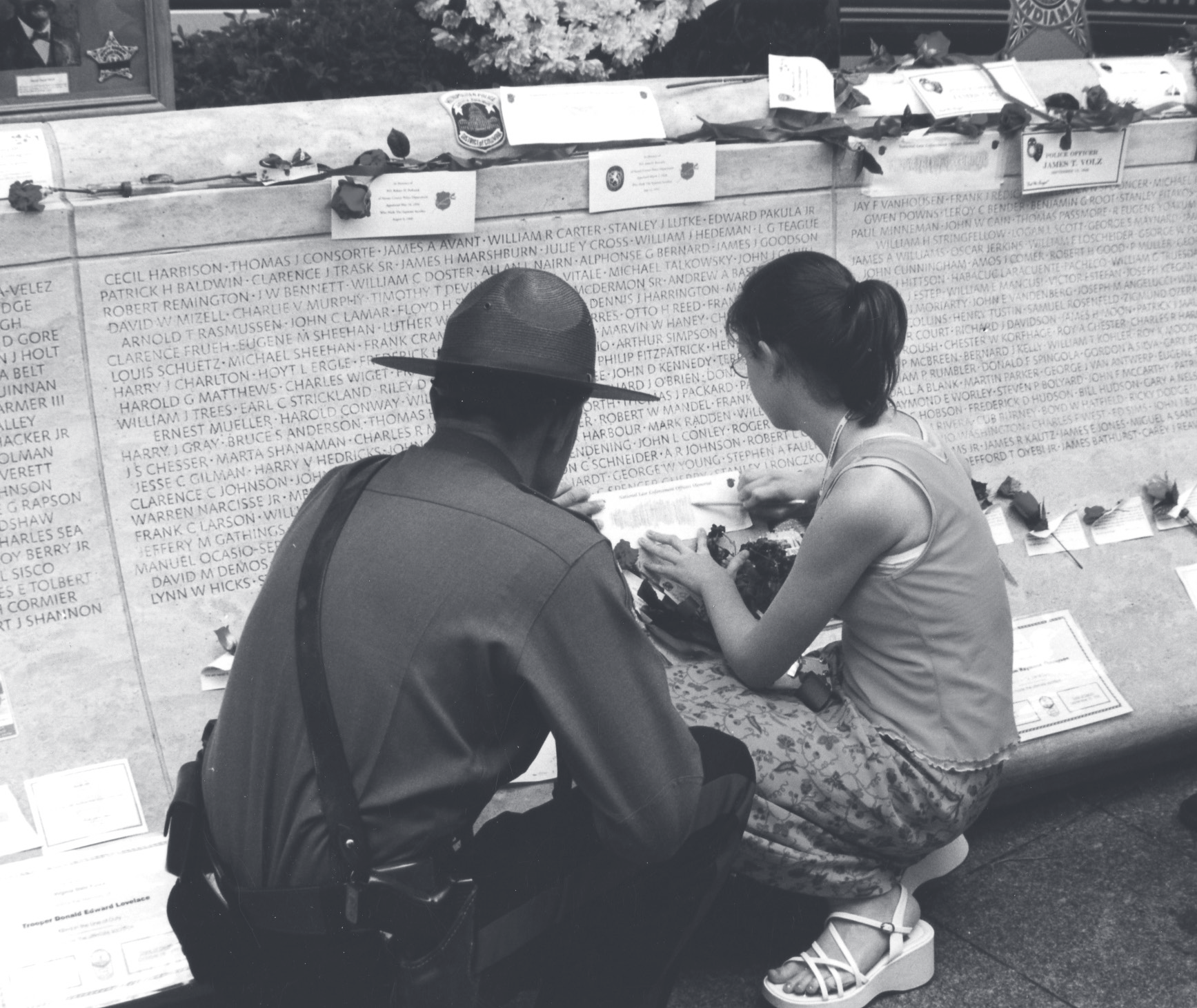 A uniformed trooper and a young woman kneel together placing items at a memorial wall engraved with names.