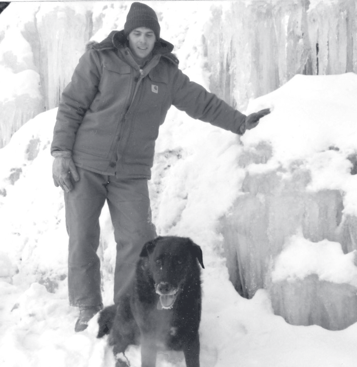 Tpr. Kolilis stands in heavy winter gear beside a black dog among large ice formations.