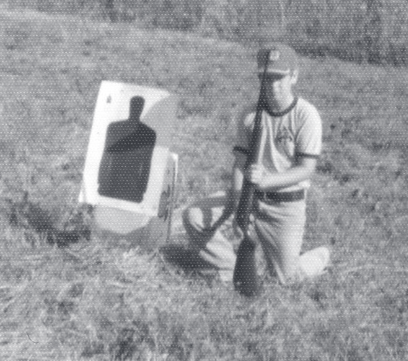 Tpr. Kolilis kneels in grass beside a paper target, holding a rifle during firearms practice.