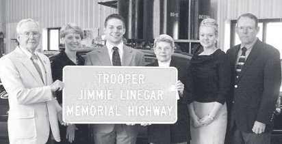 Six family members stand together holding a white highway sign reading Trooper Jimmie Linegar Memorial Highway.