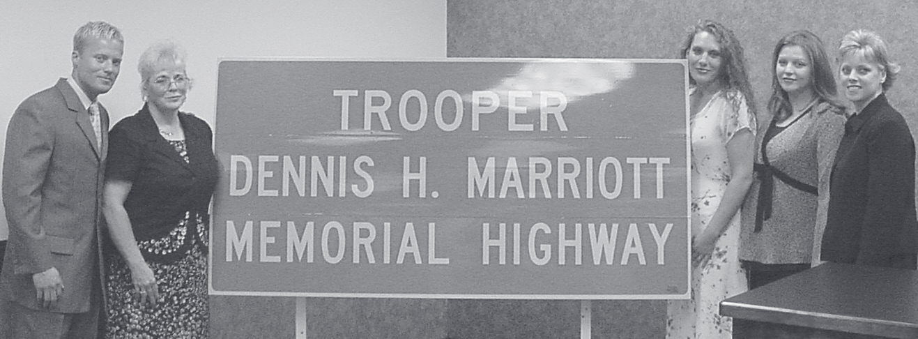 Four people stand beside a highway sign reading 'Trooper Dennis H. Marriott Memorial Highway.'