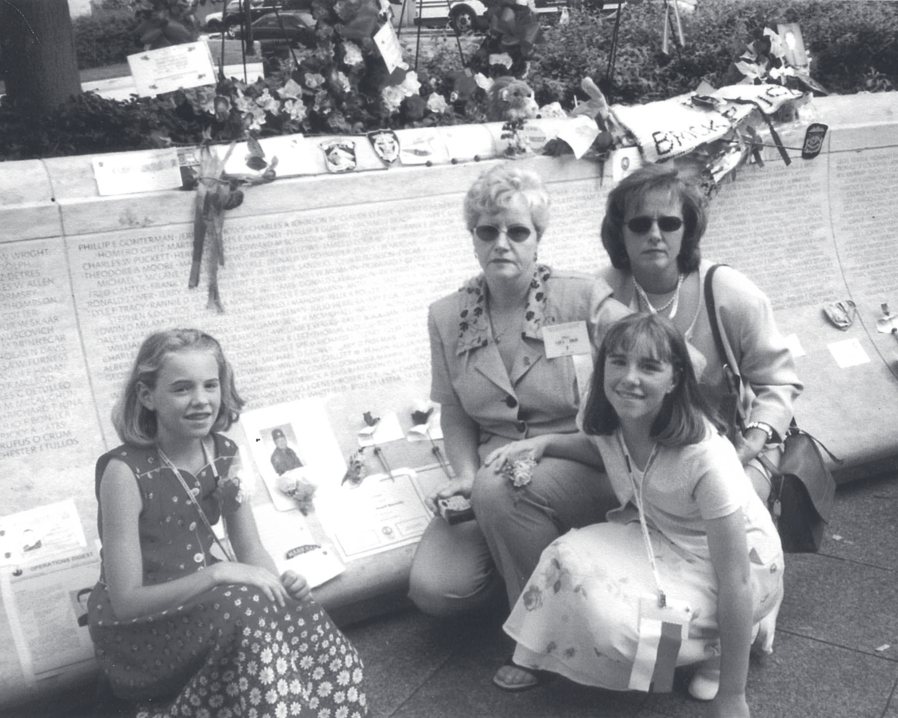 Two women and two young girls kneel beside a memorial wall with engraved names, flowers, and tribute items.