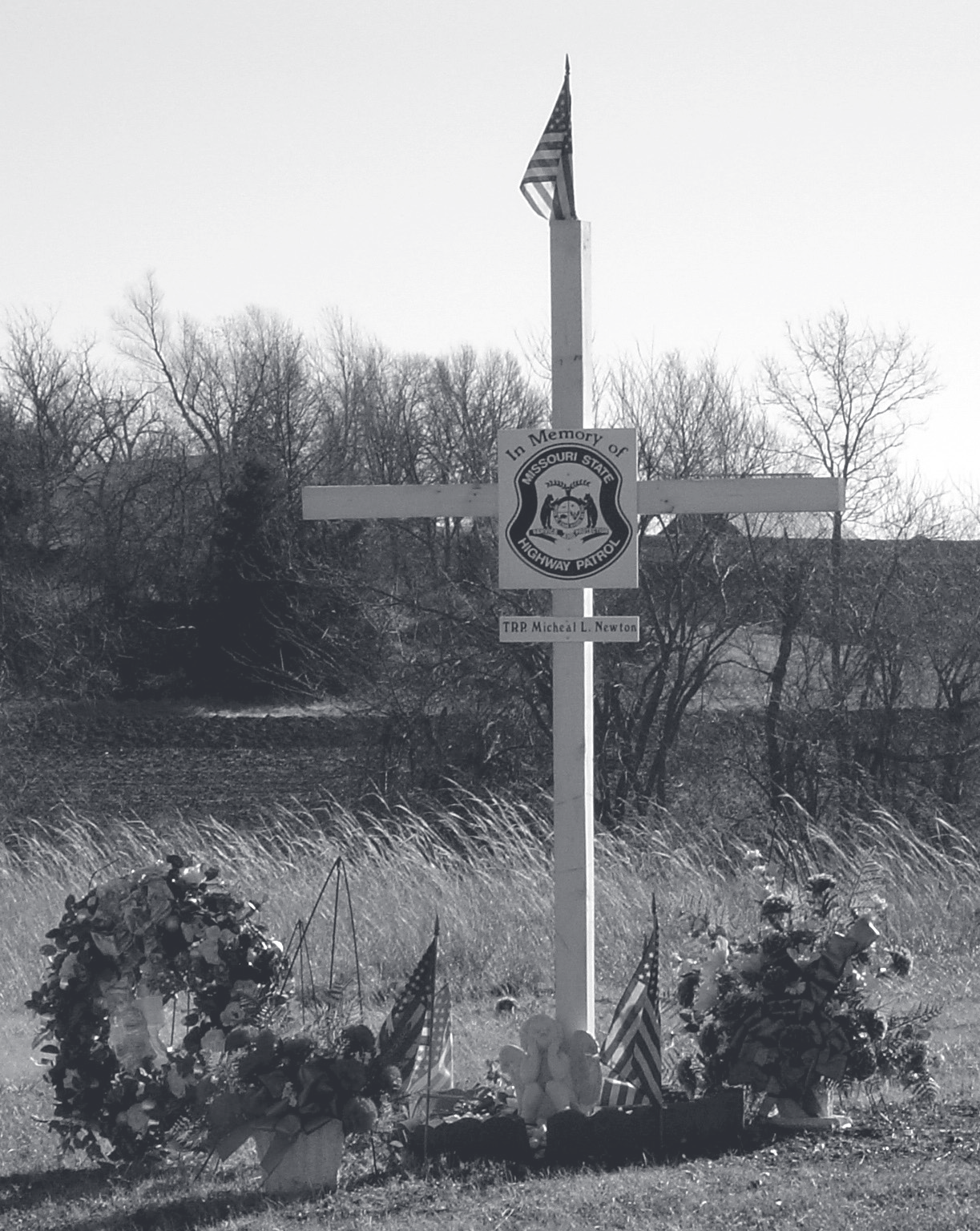Roadside white memorial cross with Patrol emblem, American flag, and floral wreaths along a grassy field.