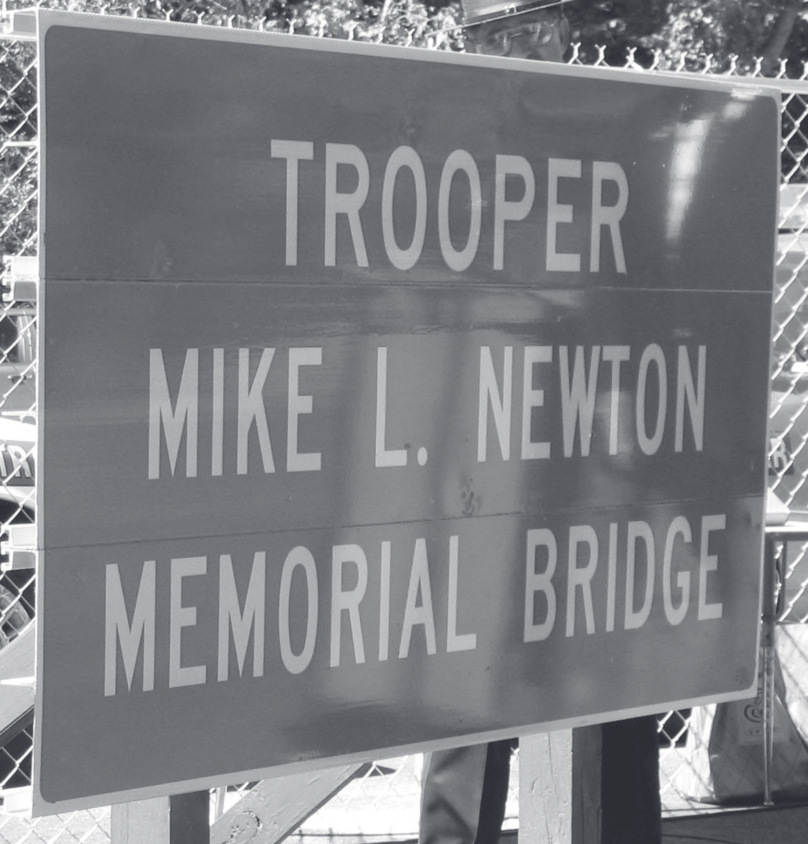 Highway sign reading Trooper Mike L. Newton Memorial Bridge mounted on a chain-link fence.
