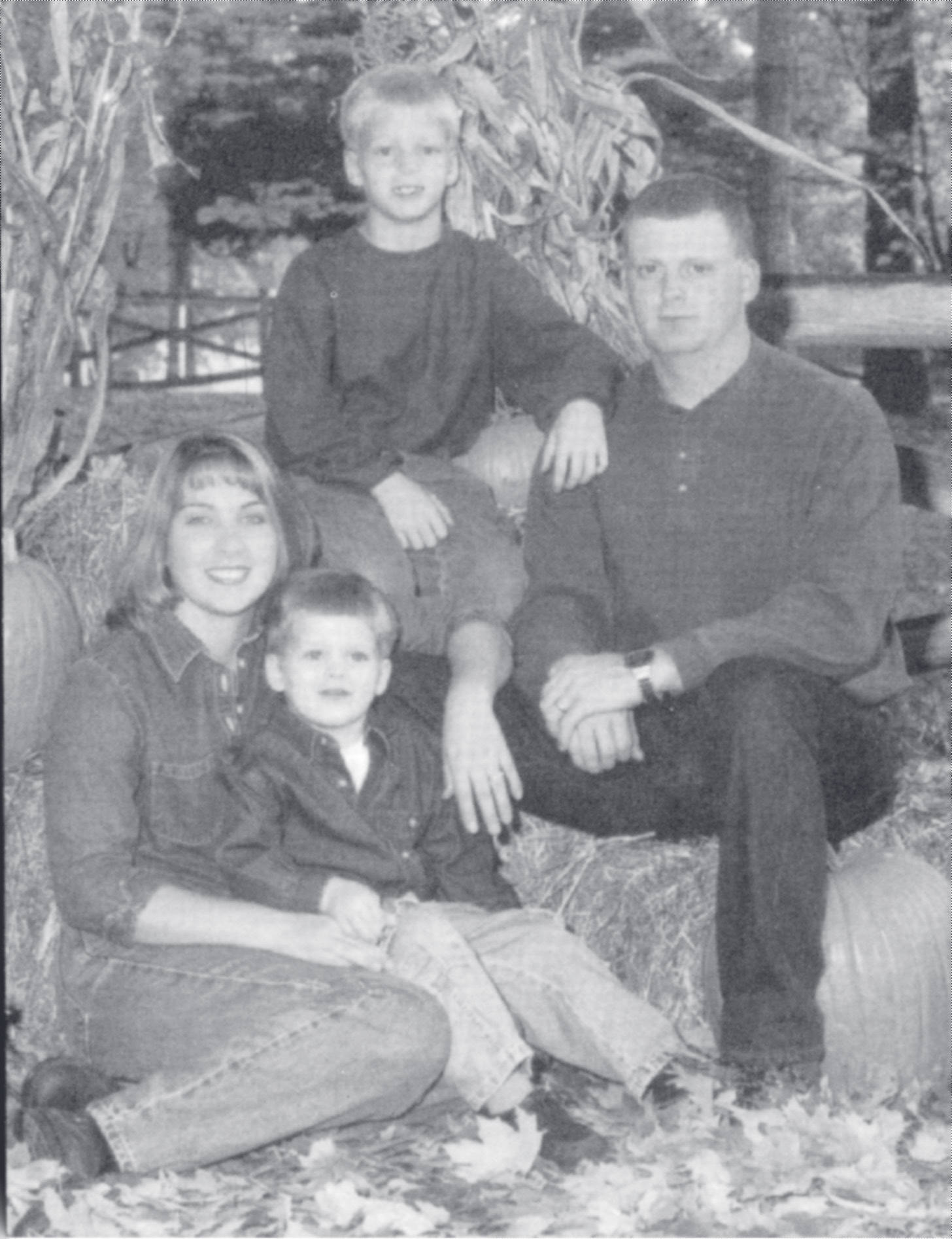 Tpr. Poynter sits beside a woman and two young boys posed on hay bales among autumn leaves for a family portrait.