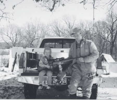 Tpr. Poynter stands beside a pickup truck holding a rifle while a small child sits on the tailgate in a rural setting.