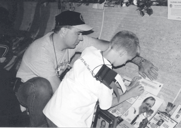 Tpr. Poynter crouches beside a young boy seated at a table signing or reading materials at an outdoor event.