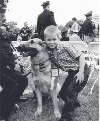 A young boy kneels with a German shepherd police dog at an outdoor event while officers and civilians stand behind them.
