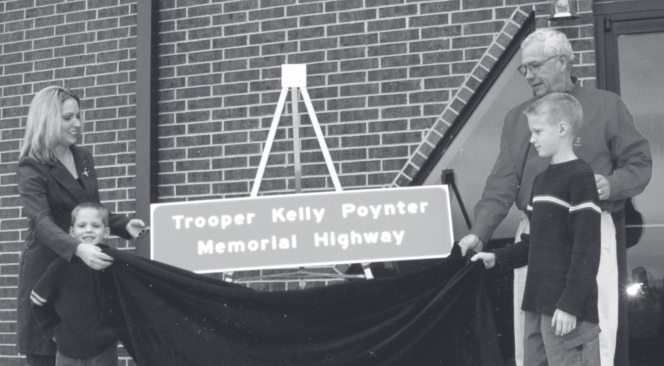 Family members and officers unveil a black-draped highway sign reading 'Trooper Kelly Poynter Memorial Highway' at a dedication ceremony.