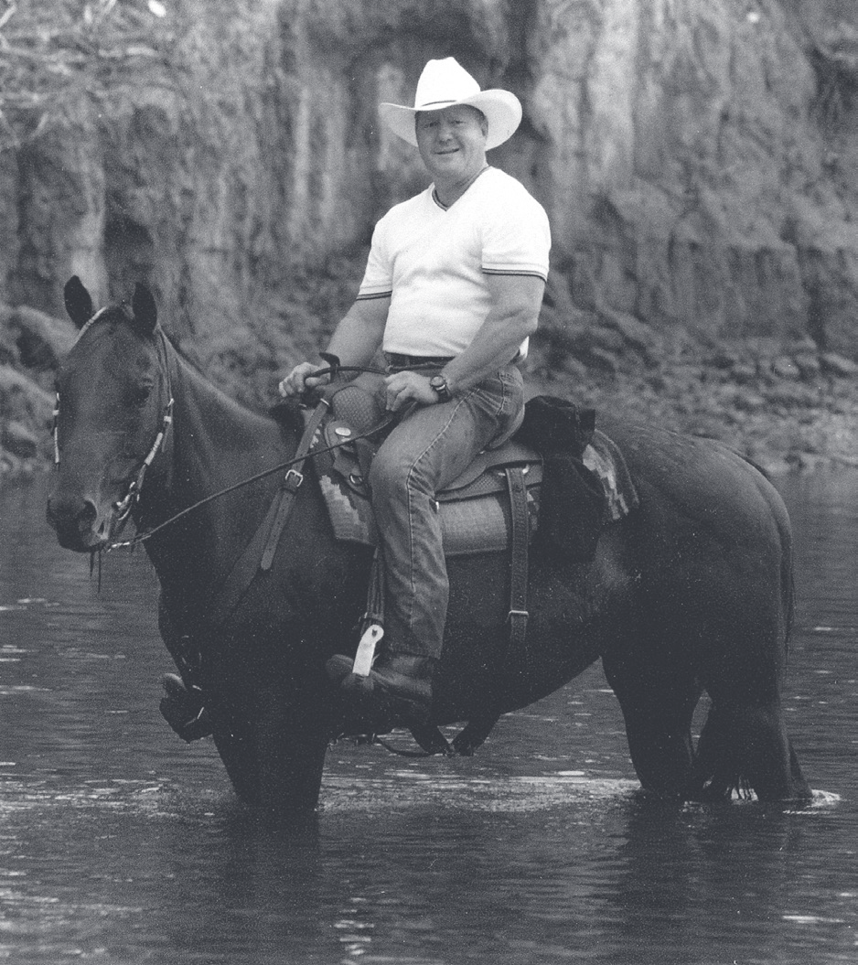 Sgt. Schuengel wears a cowboy hat while riding a dark horse through shallow water near a rocky bluff.