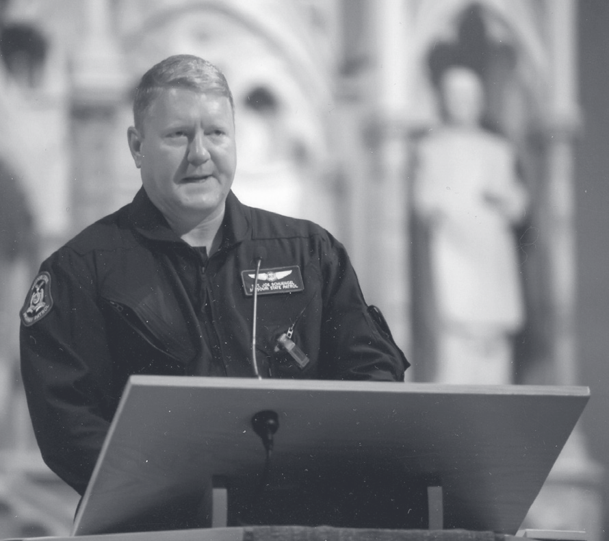 Sgt. Schuengel in uniform speaks from behind a podium at an outdoor event with statues in the background.