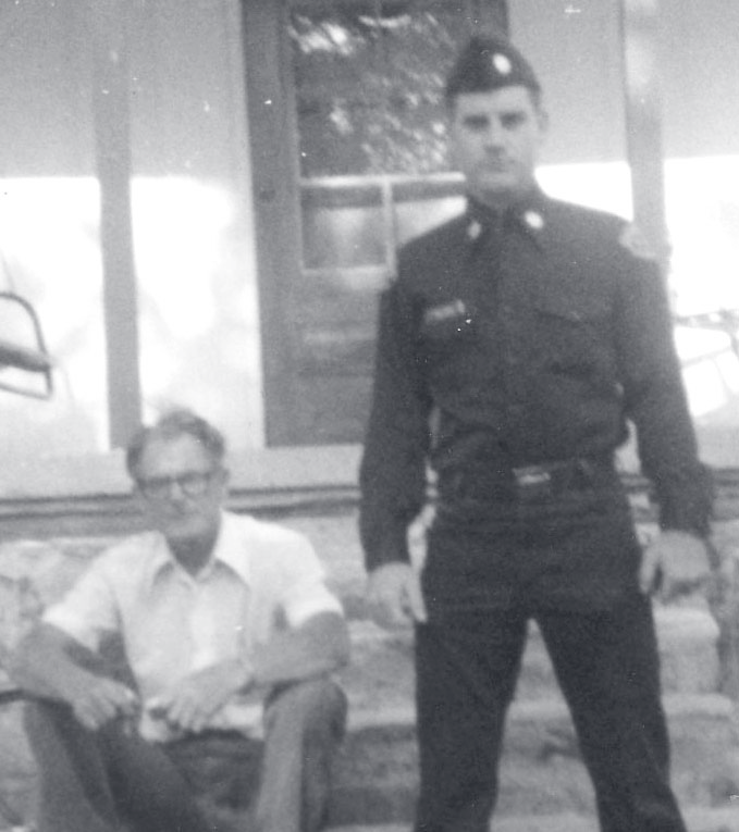 A young Sgt. Sullivan in uniform stands beside an older man seated on a porch step.