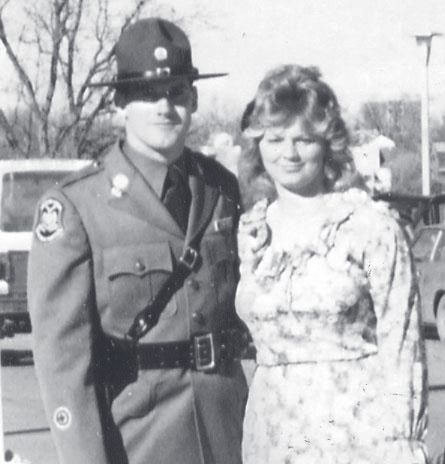 Sgt. Sullivan in dress uniform and campaign hat poses outdoors next to a woman in a patterned dress.