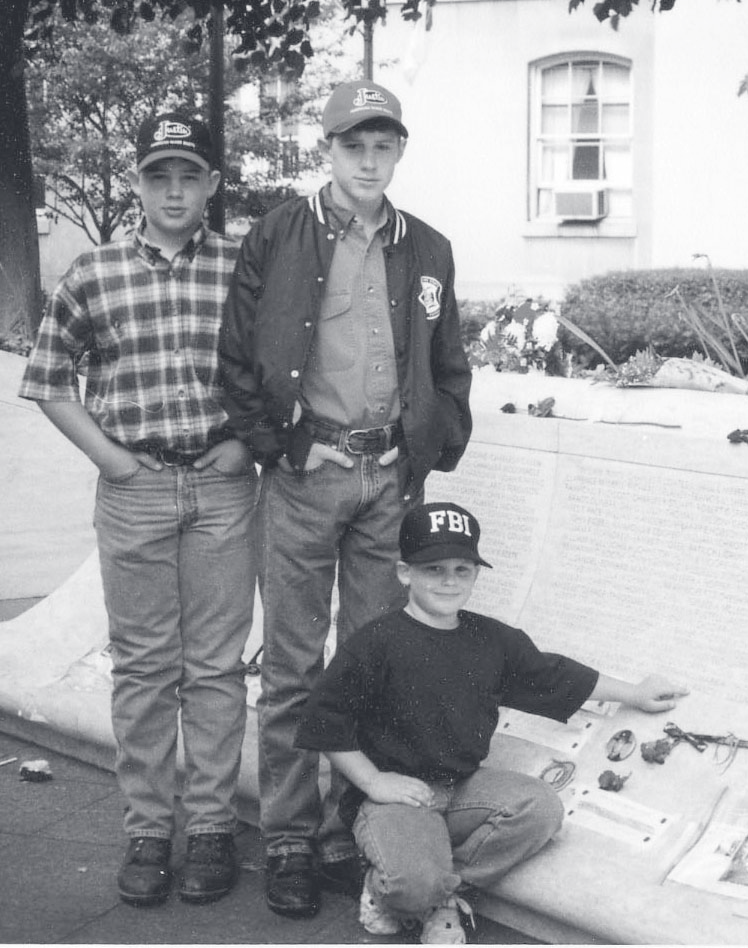 Two men in casual jackets stand behind a child in a ball cap on an outdoor walkway.