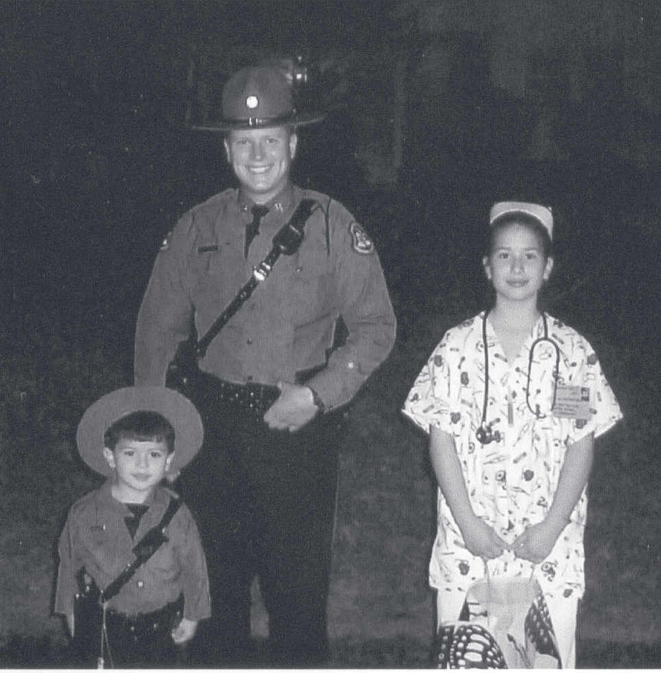 Tpr. Tatoian in uniform and campaign hat stands outdoors with two children in costume at night.