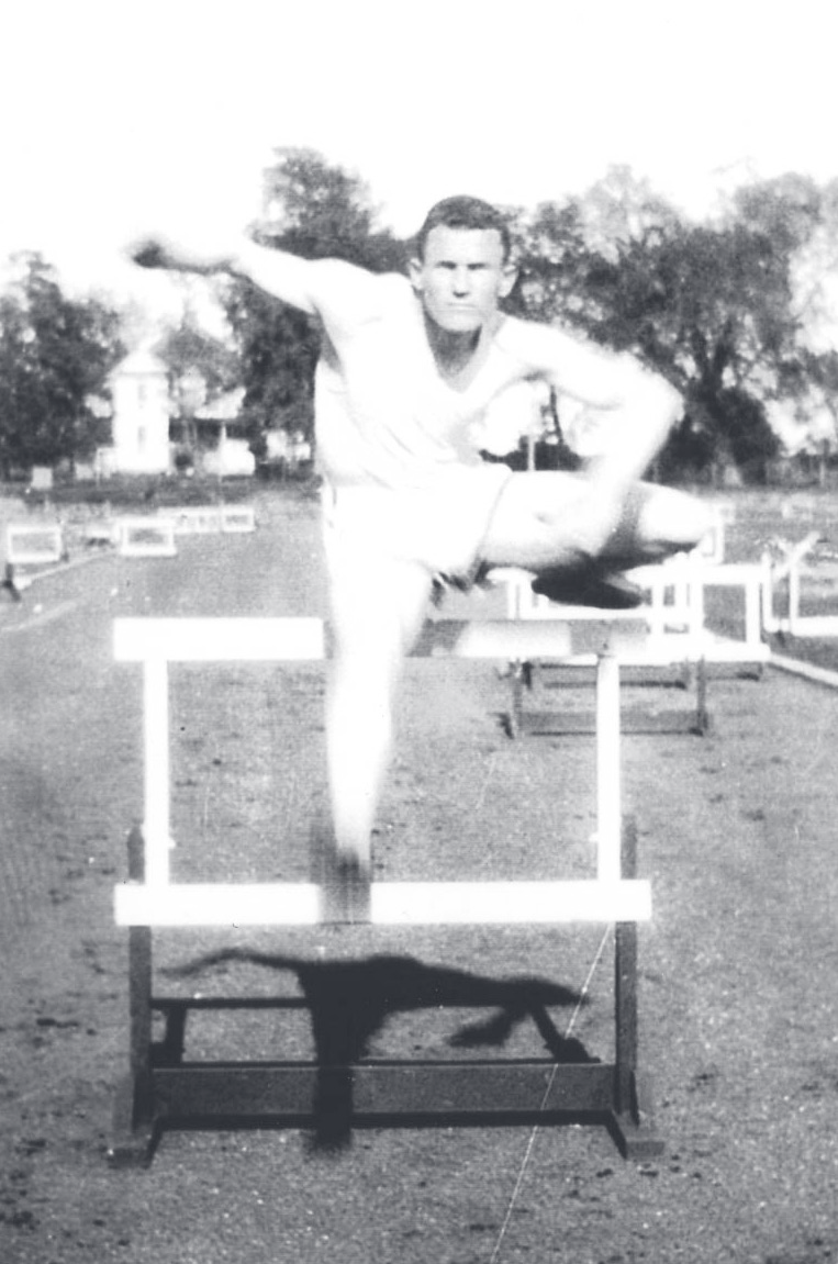 Vintage black-and-white photograph of a young man in athletic attire mid-hurdle over a track hurdle outdoors.
