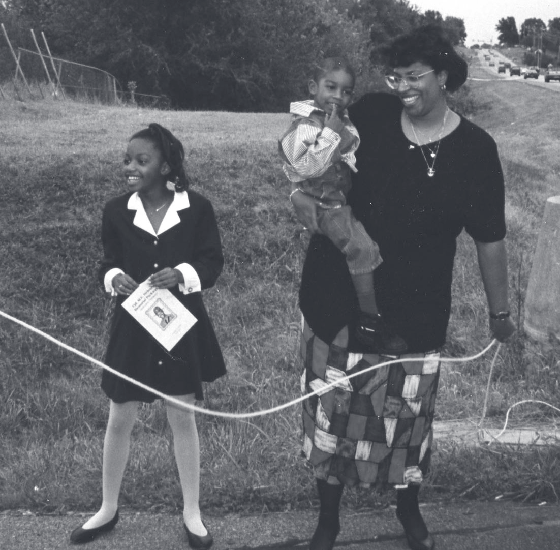 A woman and two children stand along a roadside holding a ribbon, with a highway visible in the background.