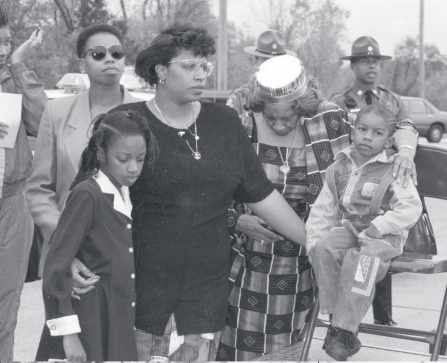 A group of family members, including children and adults, stand together at an outdoor gathering with officers nearby.